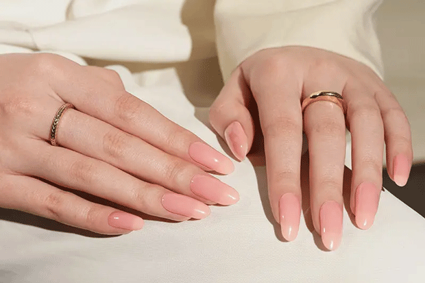 Close-up of two hands with pink nail polish strips and gold rings on a light background
