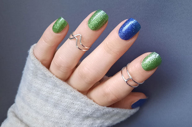 Hand with glittery blue and green nail wraps wearing multiple rings against a blurred background