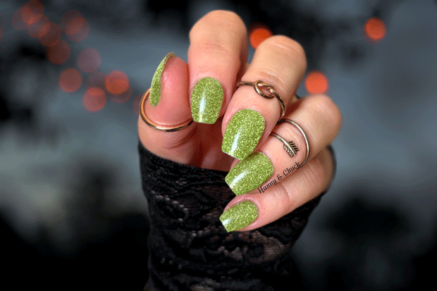 Hand with green glittery nailpolish wraps wearing black fingerless gloves against a dark background with red lights.