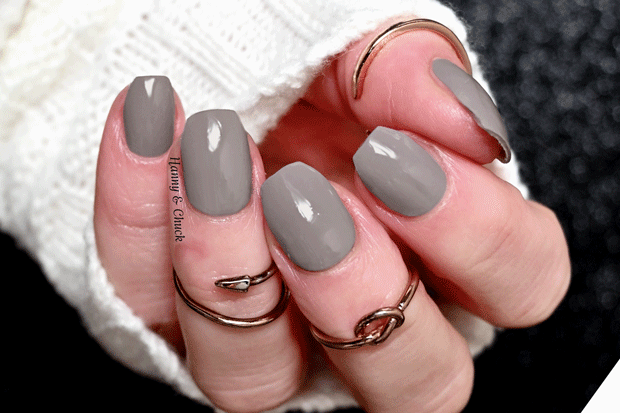 A close-up image of a hand with a fresh, dark warm gray manicure rests against a blurred, dark gray textured background.  The nails are short and neatly manicured, painted in a solid, glossy shade. 