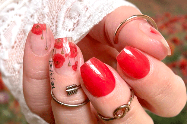 Close-up of a hand with red nail polish wraps and floral poppy designs, wearing rings, against a blurred floral background.