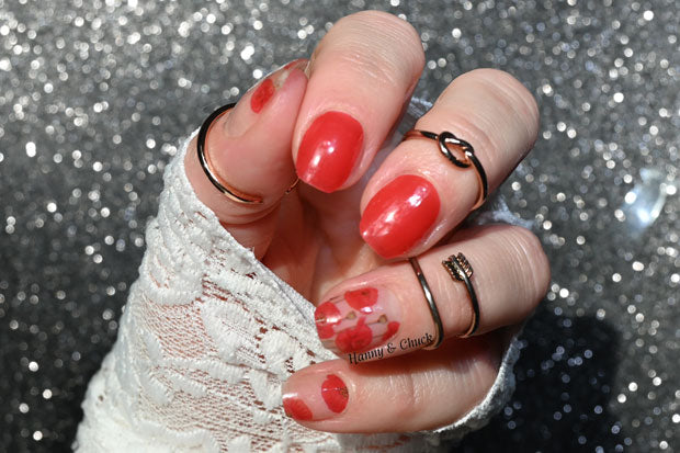 A hand with red nail polish and a sheer red nail wrap with poppy flower accents on a glittery background.